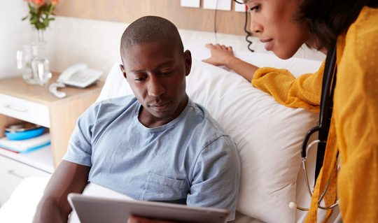 Man in hospital bed talking to staff member with tablet about paying medical bills