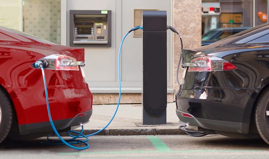 Red electric car backed into a charging station on one side and a black electric car backed into a charging station on the other side. Two opposing EVs create a visual representation of electric cars pros and cons to consider in a buyer's journey.